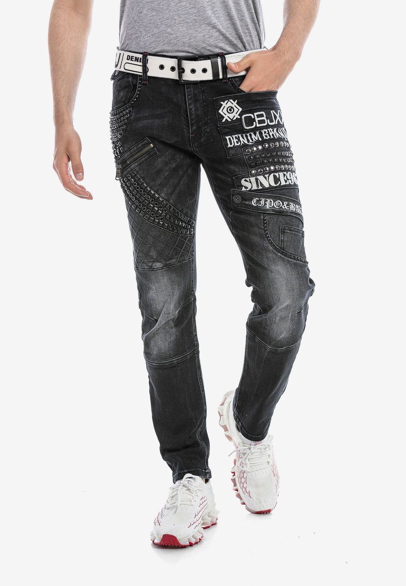Zwarte denim jeans met verschillende texturen, gestikte patronen, decoratieve studs en meerdere zakken; gedragen met witte sportschoenen.