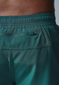 Teal atletiske shorts med elastisk talje, baglomme med lynlås, og glat stoftekstur. Designet er strømlinet og tætsiddende.