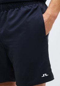 Ναυτικά μπλε αθλητικά σορτς με ελαστική μέση, χέρι στην πλαϊνή τσέπη και μικρό λευκό λογότυπο κοντά στο τελείωμα.