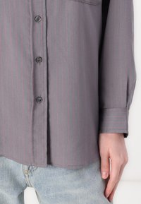 Camisa cinza de botões com riscas verticais rosa, apresentando um bolso e mangas enroladas, combinada com jeans de denim azul claro.