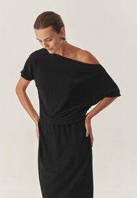 Rochie neagră, cu umerii descoperiți, cu o croială relaxată, un bust drapat și brâu elastic. Textură netedă cu o strălucire subtilă, asociată cu cercei îndrăzneți.