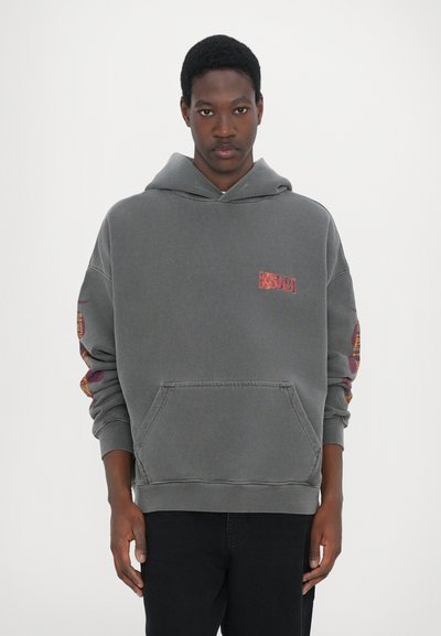 Sweat à capuche oversize gris avec poche kangourou, logo brodé sur la poitrine et accents colorés à motifs sur les manches. Matière en mélange de coton doux.