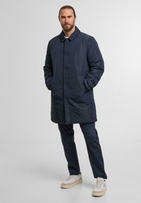 Marinefarbener Daunenmantel mit Knopfleiste, seitlichen Taschen und Kragen, kombiniert mit dunklen Hosen und hellen Sneakers.
