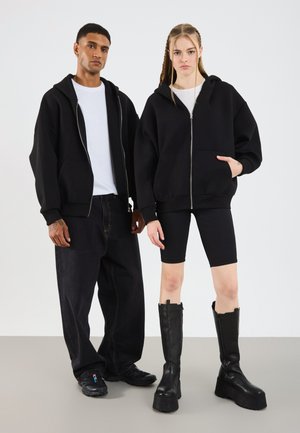 To modeller har på seg oversized svarte zip-hetter. Den mannlige modellen kombinerer sin med løse svarte jeans og sneakers; den kvinnelige modellen har på seg svarte sykkelshorts og plattformstøvler.