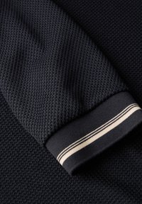 Marineblau gestrickter Stoff mit strukturierten Oberflächen. Der Bündchenbereich hat ein festes Band mit zwei beigen Streifen. Nahaufnahme des Ärmels.