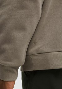 Grauer Sweatshirt mit strukturierter Wolle und Rippbündchen, mit einem kleinen Logo-Patch, das nahe dem Saum gestickt ist.