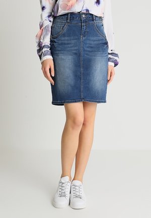 Mujer con una falda vaquera azul hasta la rodilla, blusa floral de manga larga y zapatillas blancas con cordones, de pie con las piernas cruzadas.