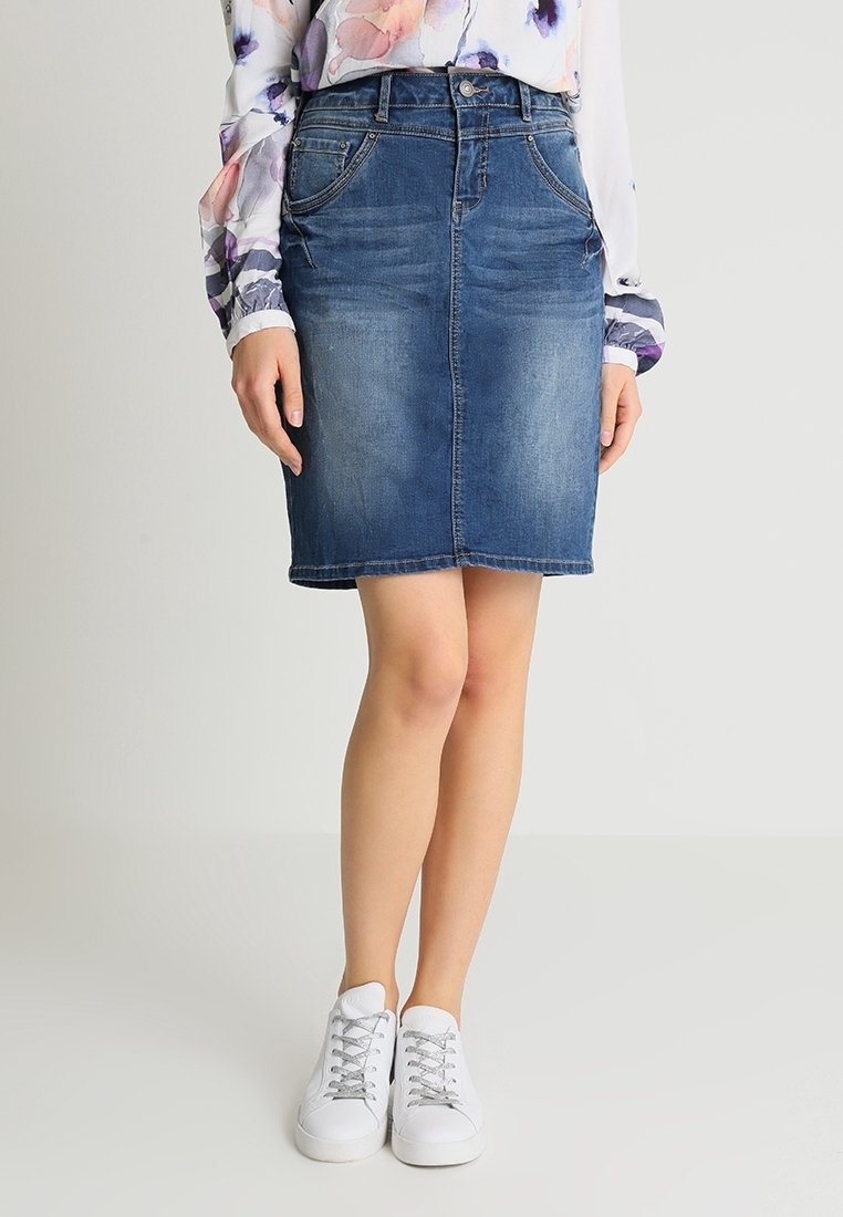 Femme portant une jupe en jean bleue longueur genou, un chemisier fleuri à manches longues et des baskets blanches à lacets, debout les jambes croisées.