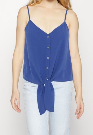 Kvinde iført en ærmeløs blå bluse med knapper foran og bindebånd foran, kombineret med lyseblå jeans.