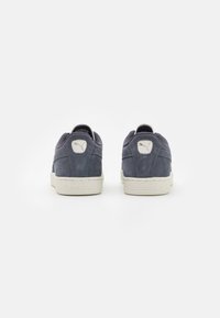 Suede modro-črne sneakers s belo gumijasto podplatom, ki imajo majhen logotip na peti in kontrastne teksturirane detajle.
