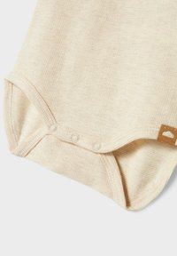 Body en coton beige présentant une texture côtelée, des fermetures par pression en bas, et une étiquette beige avec un motif de nuage sur le côté.