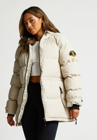 Béžová nafukovacia bunda so zipsom vpredu, s logom na rukáve a elastickými manžetami. Má prešívanú textúru a dve bočné vrecká na zips.