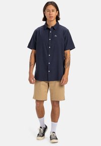 Camisa de botones de manga corta navy con un logo en el pecho, combinada con pantalones cortos caqui y zapatillas negras, usadas con calcetines blancos.