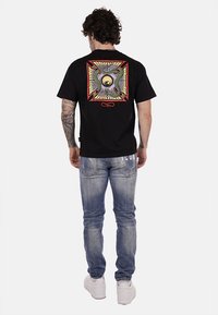 T-shirt in cotone nero con una grafica colorata di un occhio e serpenti sul retro. Abbinato a jeans di denim blu chiaro e sneakers bianche.