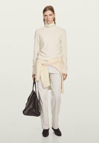 Maglione a collo alto crema, cintura avvolgente beige chiaro in vita, pantaloni bianchi su misura, borsa nera con texture e scarpe con tacco scuro. Design minimalista.