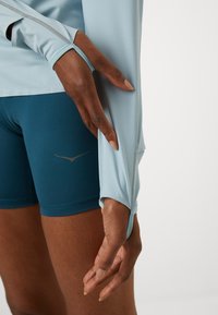 Light blue long-sleeve athletic top with thumbholes, paired with teal shorts featuring a subtle logo and smooth fabric texture.