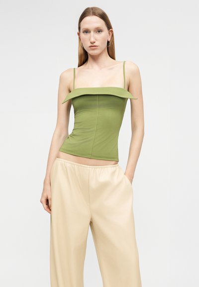 Γυναίκα που φοράει ένα πράσινο κορμάκι χωρίς τιράντες με διπλωμένο ντεκολτέ και μπεζ παντελόνι με φαρδιά μπατζάκια, όρθια με το ένα χέρι στην τσέπη της.