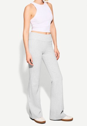 Femme portant un crop top blanc sans manches, un pantalon large gris clair et des baskets blanches à semelles marron, debout devant un fond uni.
