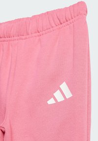 Un pantalon de survêtement rose en tissu doux avec une taille élastique et un logo Adidas blanc sur la cuisse gauche. Texture lisse et design décontracté.