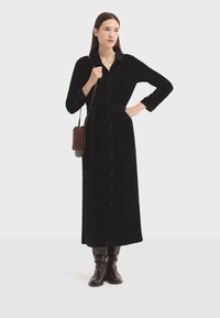 Donna in piedi con un lungo vestito nero con bottoni, una cintura, stivali marroni e una borsa a spalla marrone su uno sfondo bianco semplice.