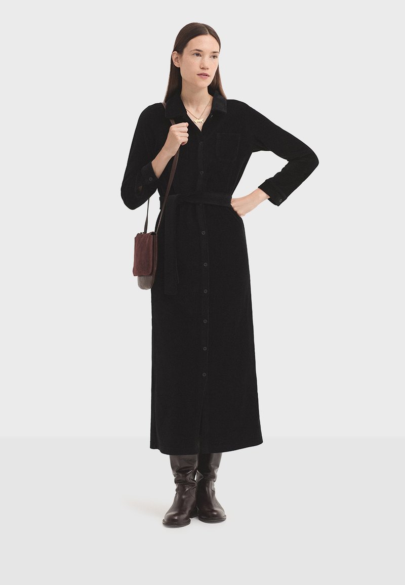 Donna in piedi con un lungo vestito nero con bottoni, una cintura, stivali marroni e una borsa a spalla marrone su uno sfondo bianco semplice.