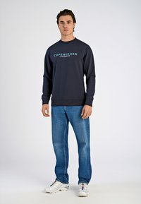 Marineblaues Sweatshirt mit dem Text "Copenhagen Lindbergh", Rundhalsausschnitt, gerippte Ärmelbündchen, kombiniert mit blauen Jeans und weißen Turnschuhen, auf einem einfarbigen Hintergrund.