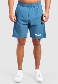 Hombre con shorts deportivos azules con un logo blanco en la pierna izquierda y zapatillas blancas, de pie contra un fondo liso.