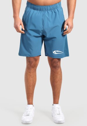 Mann trägt blaue Sportshorts mit einem weißen Logo am linken Bein und weißen Turnschuhen, steht vor einem einfarbigen Hintergrund.