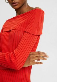 Haut en tricot côtelé rouge avec un design dénudé d'épaules, mettant en avant un détail plissé le long du col et des manches longues. Texture lisse.