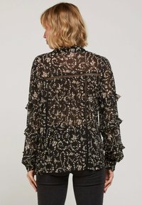 Blouse noire à motif floral avec un tissu transparent, des manches volantées et un col haut. Présente un design à motifs dans des tons crèmes atténués.