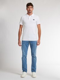 Bílá polokošile s krátkými rukávy a límečkem, v kombinaci se světle modrými džínami a bílými teniskami. Obsahuje malé logo na hrudi.