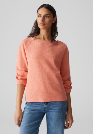 Maglione in tessuto corallo con scollatura ampia e maniche arrotolate, abbinato a jeans blu. Il tessuto ha un aspetto morbido e accogliente.