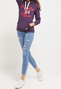 Sudadera morada con estampado "Sunday Japan 54", capucha con cordones, bolsillo delantero. Combinada con jeans desgastados de color azul claro y zapatillas blancas.