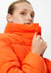 Orange pufferjakke med høj krave, lynlås og trykknaplukninger. Har quiltet tekstur og let glans i stoffet.