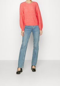 Korallfarbener, strukturierten Strickpullover mit langen, puffigen Ärmeln, kombiniert mit hellblauen, geraden Jeans und schwarzen Plateausandalen.
