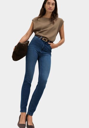 Kvinde iført brun ærmeløs top og højttaljet blå stramme jeans, der holder en mørkebrun håndtaske og har mørkebrun hæle på.