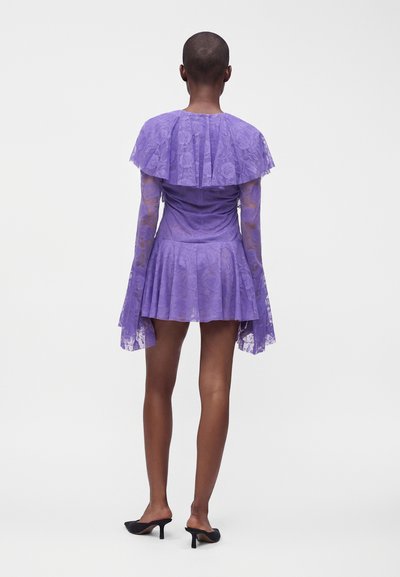 Personne portant une robe en dentelle violette à manches longues et jupe évasée, debout de dos, portant des chaussures à talons noires.