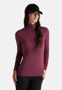 Maglione a maniche lunghe con collo alto in tessuto viola profondo, dalla superficie liscia. Abbinato a un berretto nero con un logo discreto sul fronte.