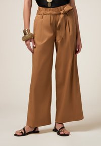 Pantaloni a vita alta e larghi in tessuto marrone chiaro, con vita legata e tasche laterali, abbinati a sandali flat neri.