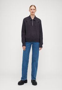 Mørk grå strikkegenser med stor krage og halv glidelås, kombinert med lyseblå rette jeans og svarte platå sko.