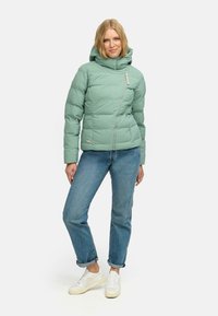 Mintgroene pufferjack met een hoge kraag, ritssluiting en zijzakken, gedragen over lichtblauwe jeans en witte sneakers.