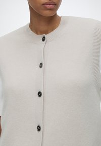 Femme portant un cardigan beige clair à manches courtes avec un col côtelé et quatre boutons noirs sur le devant, montrée du cou jusqu'au milieu du torse.
