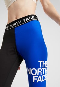 Legginsy z wysokim stanem w kolorze czarnym i niebieskim, z bocznym rozcięciem oraz białym logo po boku i szerokim elastycznym pasem.