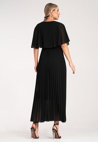 Czarna, plisowana suknia maxi z przezroczystą pelerynką na górze. Charakteryzuje się zwiewnym fasonem i półprzezroczystą teksturą. Wykończona przezroczystymi pantoflami na obcasie.