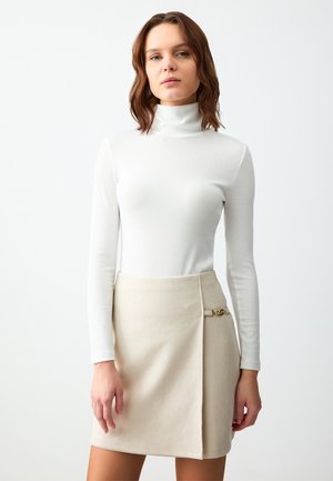 Körperbetontes weißes, geripptes Rollkragenshirt mit langen Ärmeln, kombiniert mit einem beigen Wickelrock, der mit einem goldenen Akzent am Bund versehen ist.