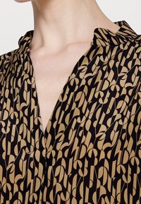 Black blouse with a v-neckline, featuring a bold gold abstract pattern. Smooth texture and lightweight fabric. Short collar detail.