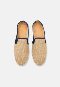 Des chaussures légères beige clair à enfiler, avec une tige en fibres naturelles tissées, des accents élastiques navy et une doublure intérieure lisse, comportant des perforations pour la respirabilité.