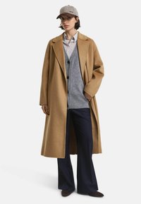 Kamelhaar-Wollmantel mit taillierter Passform, grauer Strickweste und navyfarbener weiter Hose. Ergänzt durch eine graue Kappe mit Logo.