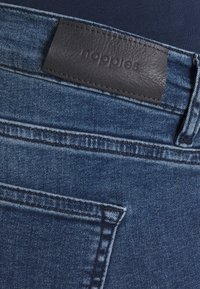 Jeans i denim med medelblå tvätt och en läderlapp på midjan som visar varumärket. Har synlig sömnad och en bakficka.