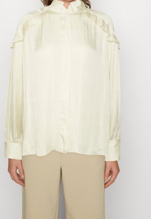 Frau trägt eine cremefarbene Satinbluse mit gerafften Schulterdetails und beige Hosen, steht vor einem schlichten Hintergrund.